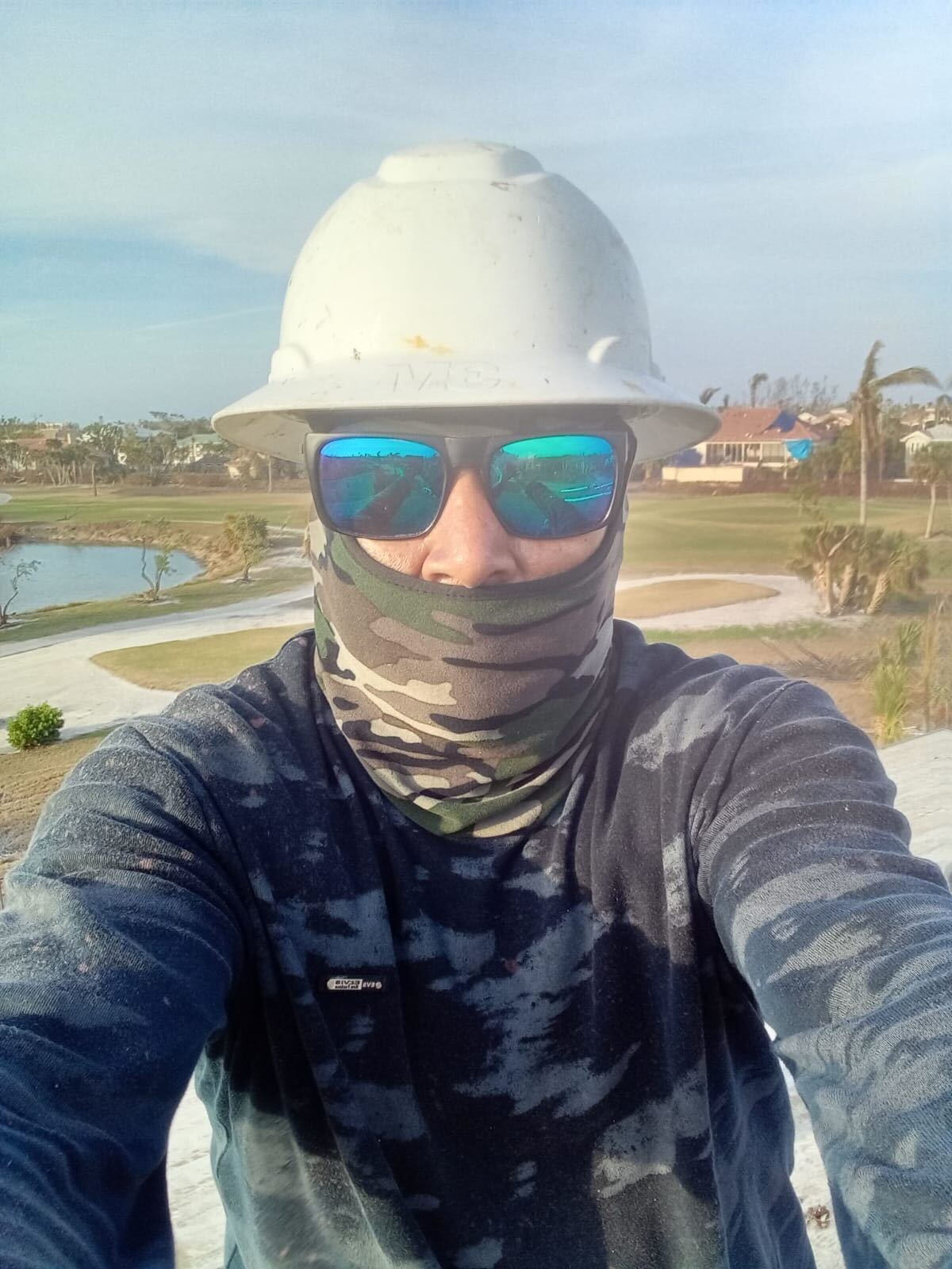 Marcos, a disaster restoration worker in Florida, poses for a selfie at work. He is wearing a white hardhat, sunglasses and a camouflage-print face covering and matching camouflage long-sleeve shirt. (Courtesy of Marcos.)