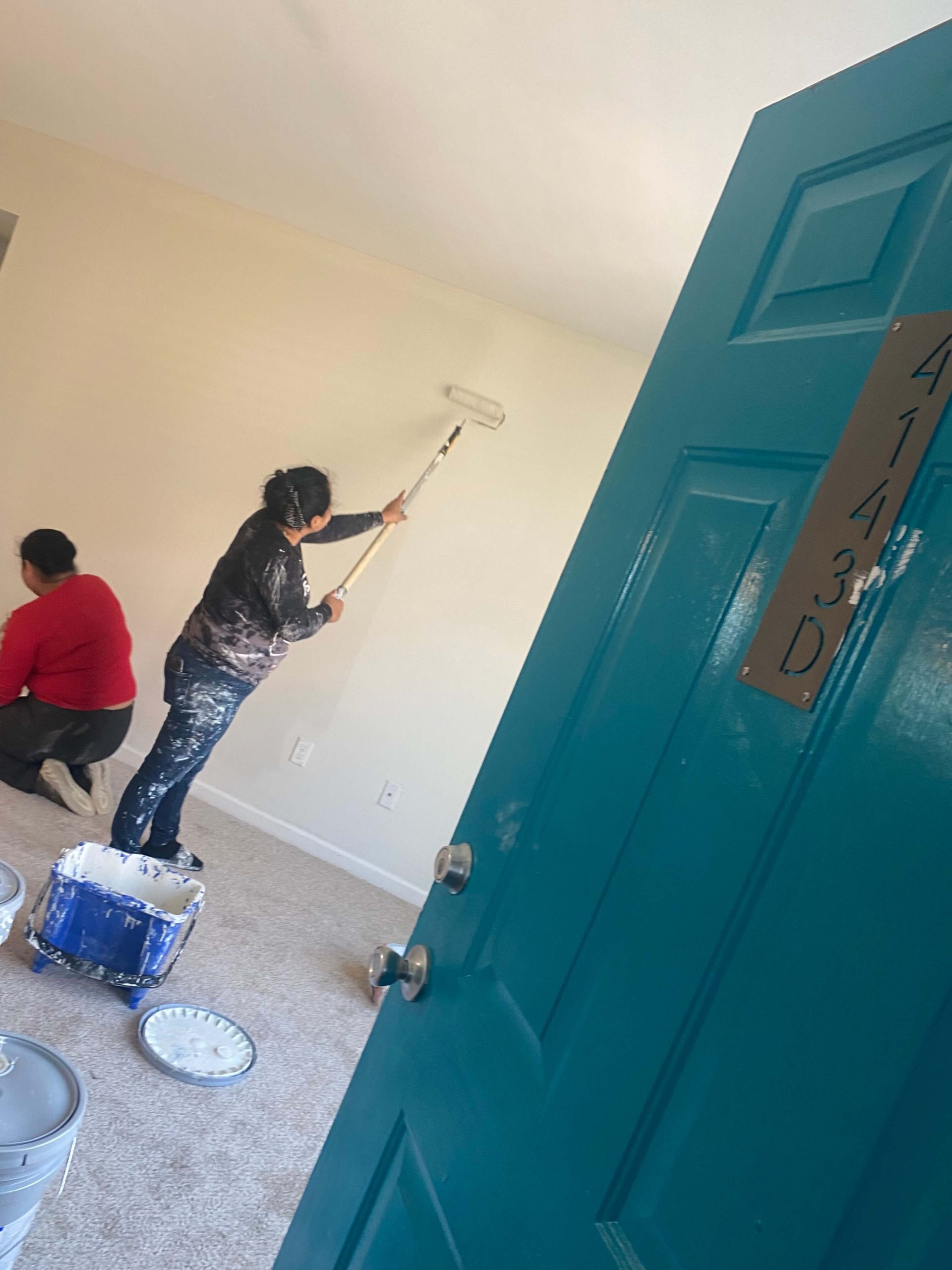 Disaster restoration worker Jenny, dressed in a black shirt and jeans, and her sister Abigail, dressed in an orange shirt and black pants, can be seen through a teal doorway, painting a vacant apartment at the Mayfair Apartment Homes, in New Orleans, with rollers and brushes, facing away from the camera. The right side of Jenny’s body is covered in splotches of white paint. (Courtesy of Jenny)