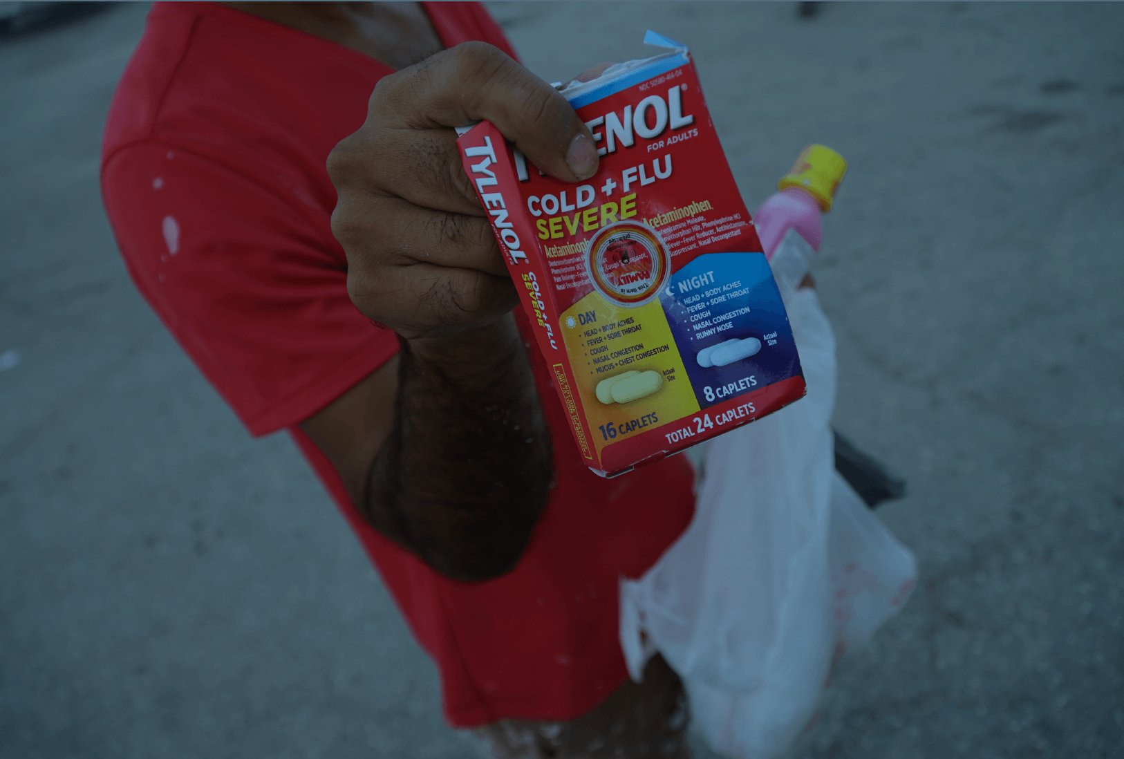 A restoration worker wearing a red shirt holds up a container of Tylenol Cold+Flu, with Pepto Bismol in his other hand. His face is not within the frame. The worker stands in a parking lot in Fort Myers where workers are picked up to clear out debris from structures destroyed by Hurricane Ian. (Jiahui Huang/Columbia Journalism Investigations)